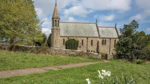A Victorian stone church with a wall around it surrounded by fields