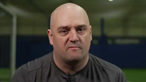 Head and shoulders shot of Brian Price. the is bald and is wearing a grey sports top. The indoor football pitch is in the background