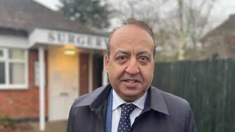 Zuffar Haq wearing a blue suit with a white striped shirt and navy tie looks at the camera.