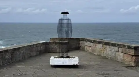 Christopher Bryceland Sculpture of soldier constructed from vertical steel plates standing on a castle wall with the sea in the background.