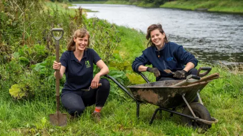 Two women with a wheelbarrow and spades at the side of a river are ready to excavate and look for archaeological items of value