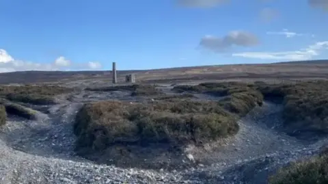 Picture of Cross Vein Mine in Foxdale. The original chimney and out-house can be seen in the distance. Around it is a flat, grey gravely surface.