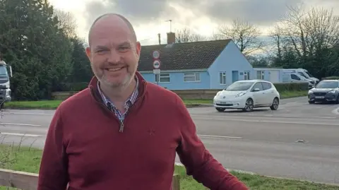 Dan Roper standing outside in front of the Crostwick Lane junction. He is looking directly at the camera and smiling. He is wearing a shirt with a red quarter zip over the top.
