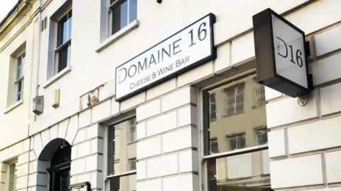 A white building with a number of windows and a black door. Two signs are on the wall, one reads Domaine 16 cheese and wine bar and the other says D16.