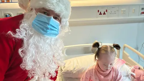 Handout Myla is sitting in a hospital bed and Santa, wearing a mask, is standing next to her looking at the camera.