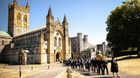 PA Media The funeral procession for Ann Widdecombe with Buckfast Abbey towering in the background against a blue sky backdrop.