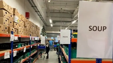 Andrew Sinclair/BBC A long aisle in a warehouse stacked with boxes and cans of food. There are signs saying "Pasta" and "Soup". A woman with a trolley is in the distance picking up cans.    