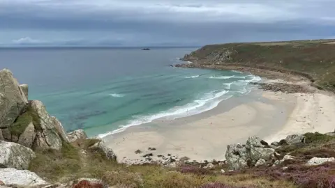 Gwynver Beach at Sennen, a large sandy beach with headland on either side. The image is taken from the top of a cliff looking down over the beach. The water is blue.