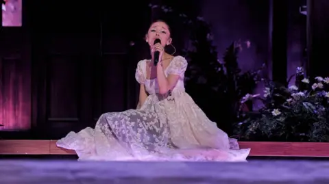 Ariana Grande sits on the stage, surrounded by flowers, as she sings on her Eternal Sunshine tour