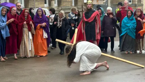 A man with long brown hair, who is wearing a white gown and a crown of thorns on his head, kneels on the floor with a large wooden cross as people gather around him on a rainy street during a re-enactment of Jesus' crucifixion