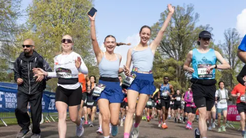 Two women in matching grey tops and blue shorts running while holding hands, with their other hands in the air. A man in a jacket, a smiling woman in a white top, and a man in a cap and blue tank top are running to their sides. dozens of other runners are seen in the background.