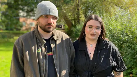 A man on the left with glasses, a grey beanie, and a short brown beard is wearing a light brown shirt open over a dark t-shirt. A woman next to him on the right also has glasses and is dressed all in black with long brown hair. They are standing outside with trees in the background.
