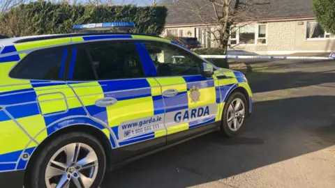 BBC A Garda car in blue and yellow in front of a Garda tape and a bungalow