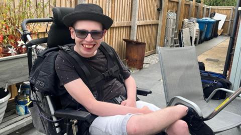James Walker in his support chair smiling at the camera wearing a black hat, shades, a t shirt and shorts sat in a garden