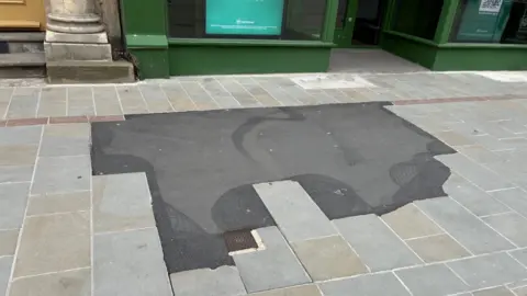 Black tarmac in place of several stone slabs on the pavement. It looks out of place compared to the clean paving stones. 