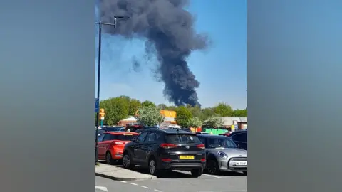 Large fire over a car park. A black column of smoke can be seen in the blue sky.