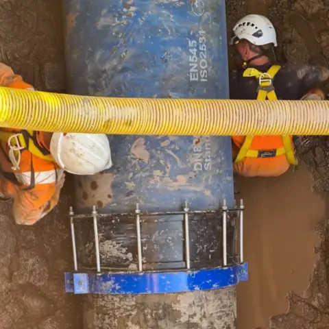 Welsh Water Two workers stand either side of a large blue water pipe, with mud surrounding them. They are both wearing white helmets and high vis orange protective clothing. A smaller yellow pipe passes over them. 