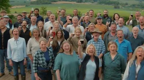 Hawkstone Farmers' Choir The choir is standing outside and looking at the camera, many toasting the picture with a pint. There are men and women with Jeremy Clarkson in the middle.