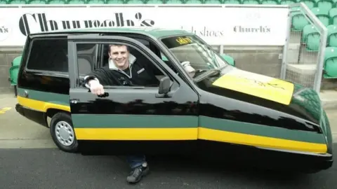 Northampton Saints Jon Sleightholme with short dark hair sitting in the driver's seat of the car which is painted in Saints colours
