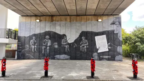 A greyscale mural painted on the wall of an underpass showing five soldiers sitting in a trench, with one writing a letter to his parents. In front of the mural are four bollards decorated with poppies.