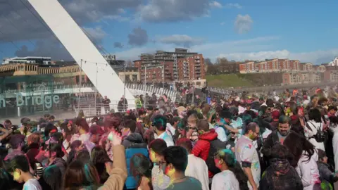 Hundreds of people covered in pain are standing near the Gateshead Millennium Bridge by the river Tyne.
