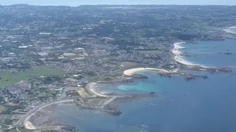 Aerial view of a coastal town with sandy beaches, rocky shoreline, turquoise bays, and patchwork fields stretching inland.