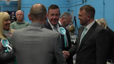 A man in the middle wearing a light blue and white rosette is looking at a man with his back to the camera. Another man on the right is smiling and shaking someone's hand, while a woman with blonde hair is smiling on the far left.