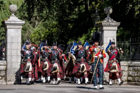 PA Media The Band of the 2nd Battalion marches through a stone gateway during a ceremonial parade. Musicians in tartan kilts and dark military uniforms play bagpipes while advancing in formation behind a drum major in a red tunic and bearskin-style hat. 