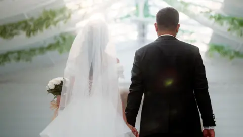 Stock photo shows the backs of a bride and groom on their wedding day, wearing a white wedding dress and black suit, the bride carries a bouquet of flowers and a marquee with flowers hanging can be seen in the background.