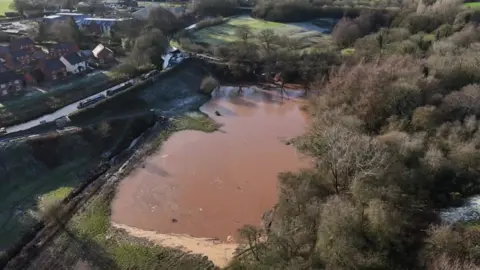 A large brown glut of water can be seen in a field next to the breached canal. Trees surround it on one side and the canal on the other side.