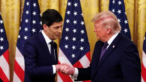 Donald Trump shakes hands with Kevin Warsh, both men wear navy suits and are flanked by US flags. 