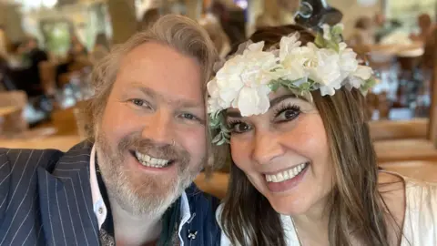 Keaton Stone Keaton and Sophia Stone smile at the camera with their faces close together. He wears a pinstripe suit and she wears a white dress and a white flower crown on her head. He has light gray hair and a beard while she has dark brown hair.