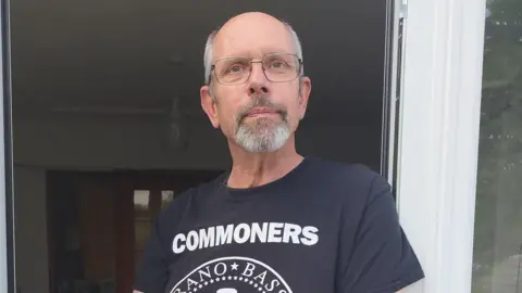 Jon Farley A man in his 60s stands by some patio doors. He is wearing a black T-shirt with white writing on and glasses.