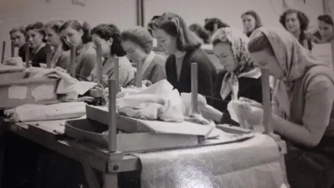 A black and white image of rows of women sitting at tables working with fabrics.