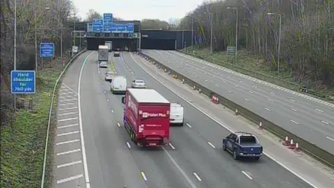 A still image from a highways camera positioned on a gantry. Below are vehicles heading away from the camera. The carriageway furthest away from the camera is empty. In the background are blue motorway signs positioned in front and above a tunnel.