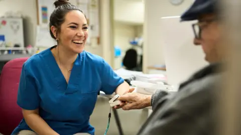A nurse wearing a blue nurse's tabard is taking an elderly man's blood pressure in a hospital. She has short dark hair in a bun and is looking directly at him with a beaming smile on her face.