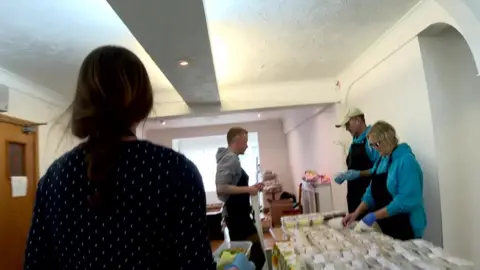 BBC Woman walks towards a table full of meals in small plastic boxes, where three other people are working, preparing to hand out the meals