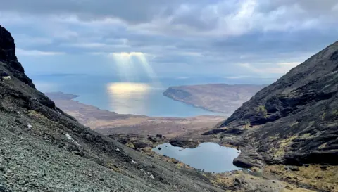 Anna Lubomirska A dramatic mountain pass opening out toward the sea. Sunbeams shine through clouds onto the water below, with rocky slopes and a small lochan in the foreground.