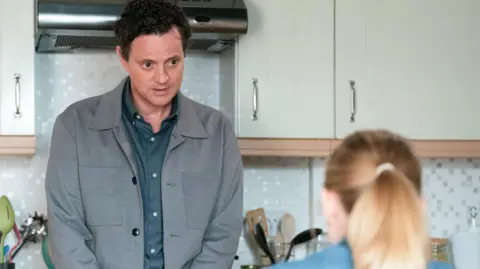 Dressed in a grey jacket and blue shirt the EastEnders character is standing in a kitchen in front of a cooker. There are jars of kitchen utensils beside him and a woman with a blonde ponytail.
