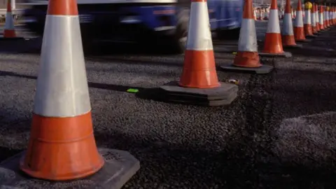 A heavy goods vehicle travelling on a road where there are orange fluorescent cones to mark part of the road.