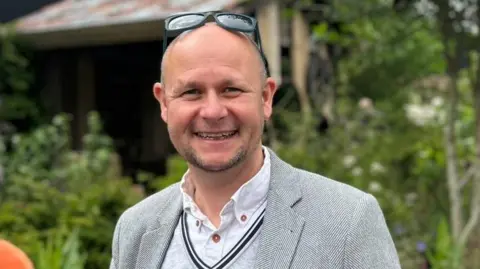 Submitted A man with a bald head and stubble beard is smiling into the camera. He has sunglasses on his head and is wearing a grey suit jacket, grey v-neck jumper over a white shirt. He is stood outside and there are trees in the background.