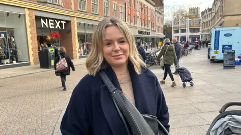 Tom Jackson/BBC Emma Taylor is in the High Street in Cambridge. She is wearing a blue coat and light brown jumper and has a buggy.