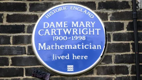 PA Media A blue plaque is on a dark brick wall. It is branded "Historic England" and says: "Dame Mary Cartwright 1900-1998 Mathematician lived here."
