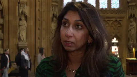 Head and shoulders picture of Suella Braverman wearing a green and black top in the central lobby of the Houses of Parliament with statues and wall carvings in the background