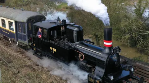 Nene Valley Railway A black steam locomotive which has steam coming out of the red and white striped funnel at the front of the machine and steam blowing along its base is running along tracks with trees behind it. A man can be seen leaning on the window in its cab and behind it is a blue and cream carriage. 