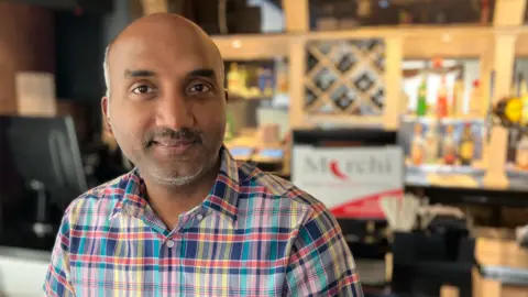 A man in a checked shirt, with short grey hair and stubble, smiles for the camera. He is stood in front of a bar, with a sign saying Mirchi over his shoulder.