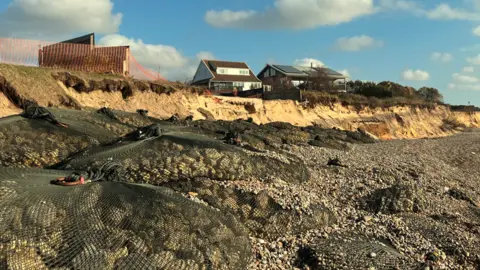 A row of large rock bags on Thorpeness beach in front of a heavily eroded coastline. In the background, there are two homes close to the edge of a low, sandy cliff. 