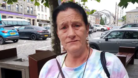 Thelma is standing in a street with cars parked behind her. She is wearing gold earrings and a tie-dye t-short. She has dark hair pulled back into a pony-tail.