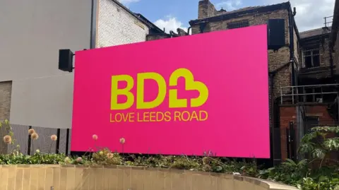 A large pink sign with the words BD3 Love Leeds Road printed on it in yellow with the 3 sign in the shape of a heart