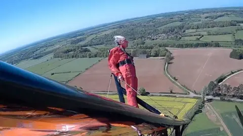 AeroSuperBatics Jenny Thurgut is strapped to the wing of an aeroplane in flight. She is wearing a red overall and standing on a black/orange wing with the wind in her face. You can see the fields below on a sunny day.